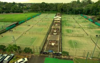 Aerial view of a tennis facility featuring multiple courts, players in action, and a central building for amenities. Lush greenery surrounds the area, with parking visible in the foreground. Ideal for tennis enthusiasts looking for a vibrant sports environment.