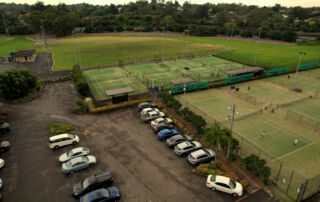 Aerial view of a tennis facility featuring multiple courts and a parking lot filled with cars, surrounded by green trees and open fields, highlighting active sports and recreational spaces.