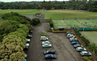 Aerial view of a parking lot filled with various cars adjacent to sports fields, including a soccer field and tennis courts, surrounded by lush greenery and trees.
